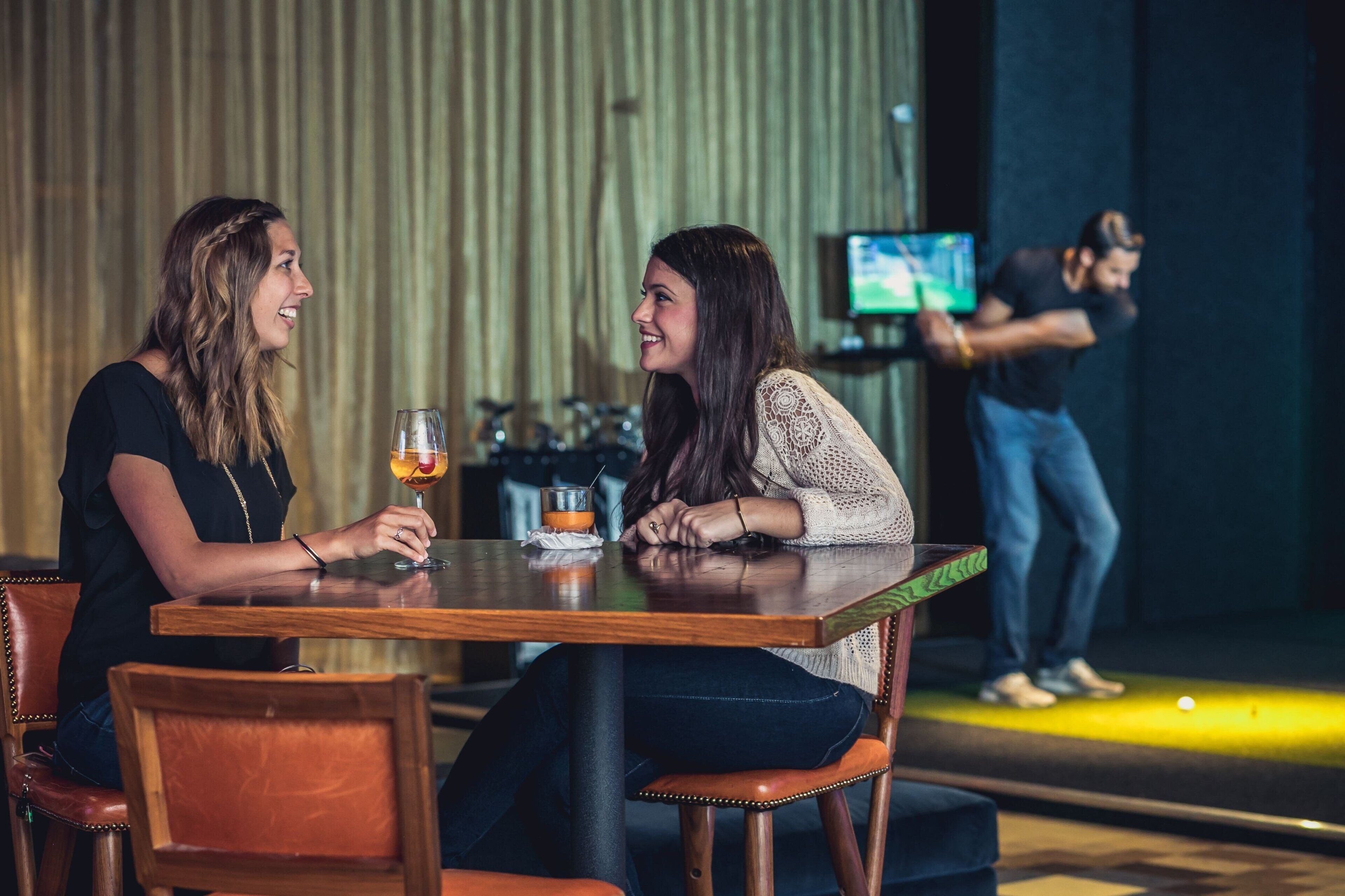 Two people seated at a wooden table with a glass of drink, in a room with a stage and a screen in the background.