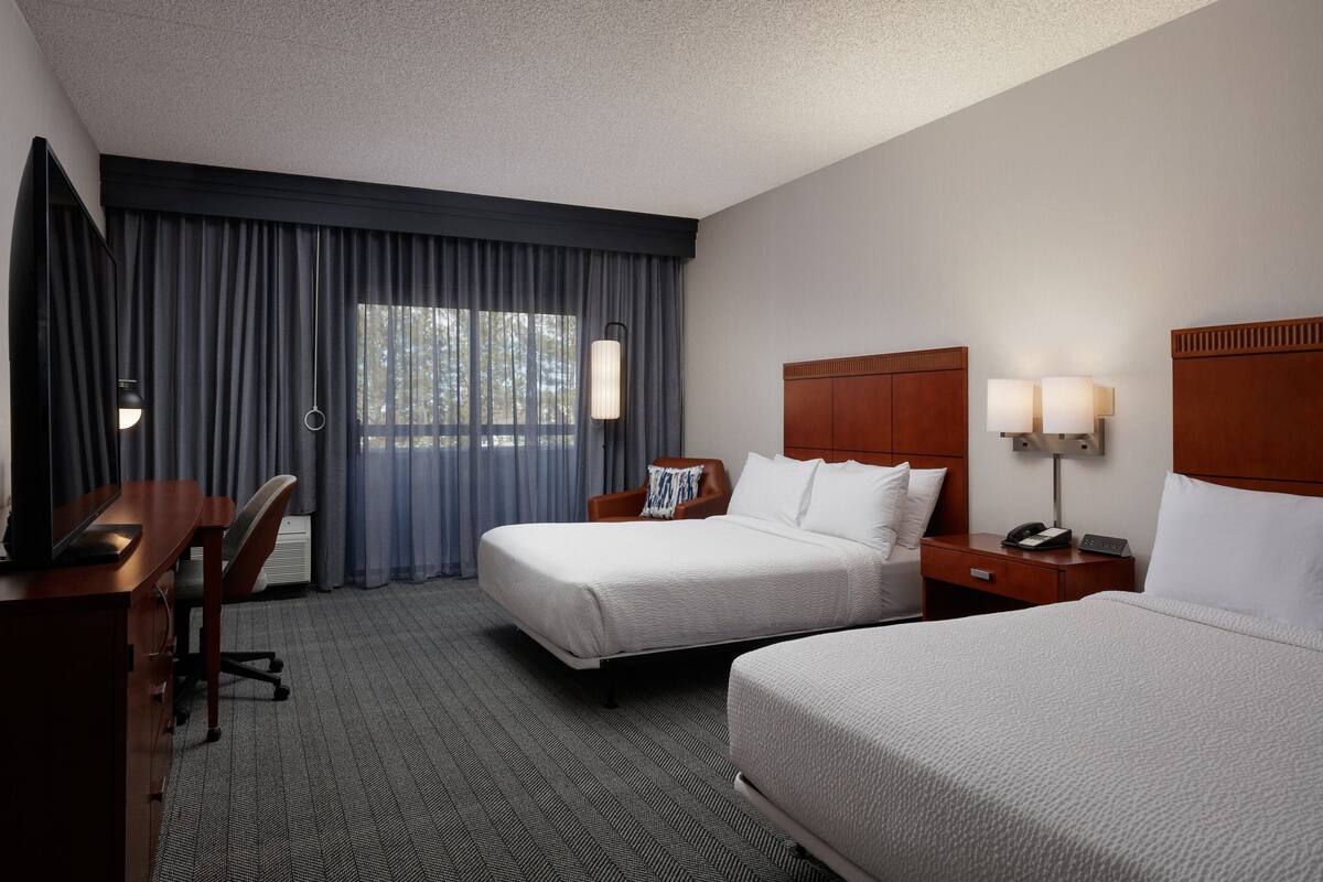 A hotel room with two beds, a desk, a chair, and a large window with curtains.