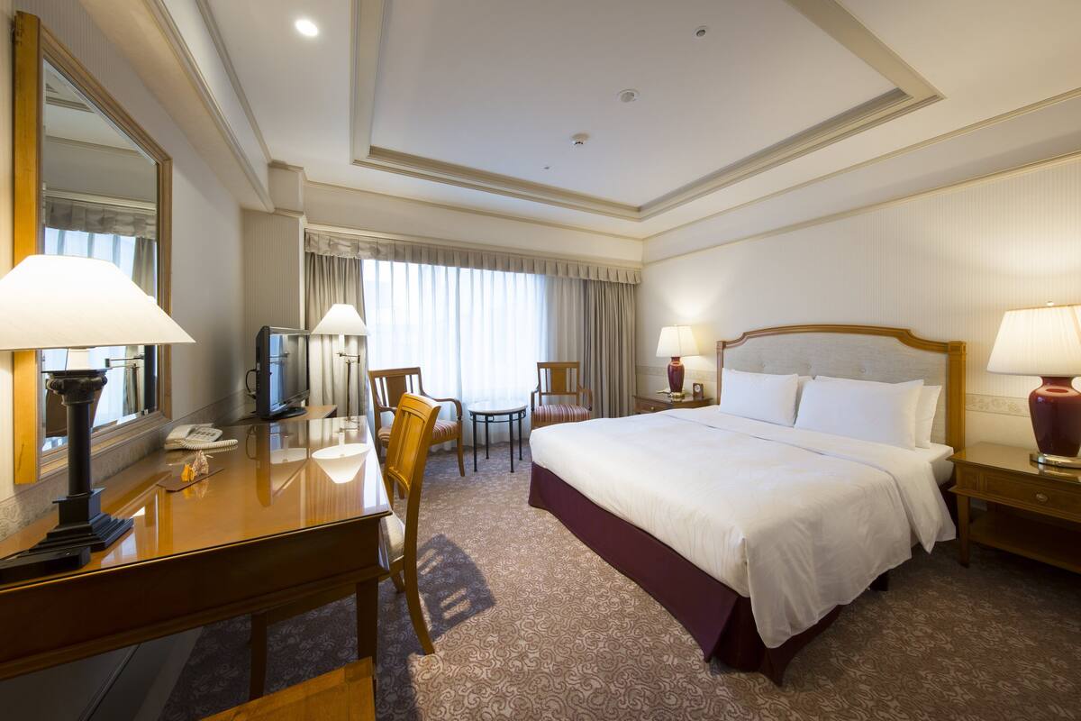 A hotel room with a large bed, a desk with a telephone, a chair, a television, and a lamp.