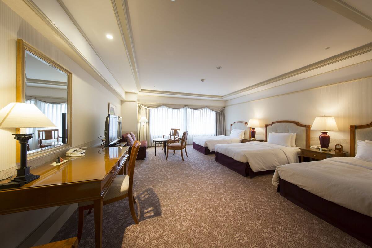 A hotel room with a desk, two beds, a mirror, and a window with curtains.