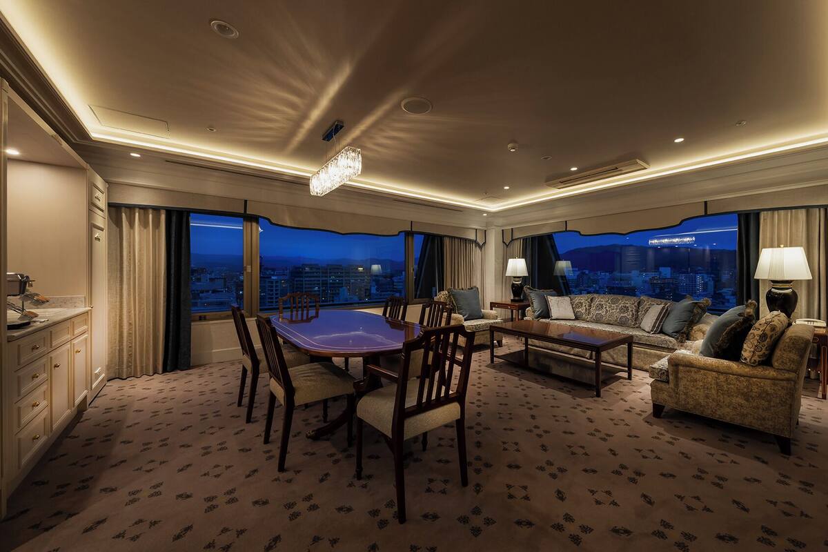 A hotel room with a dining table, sofa, and a city view.