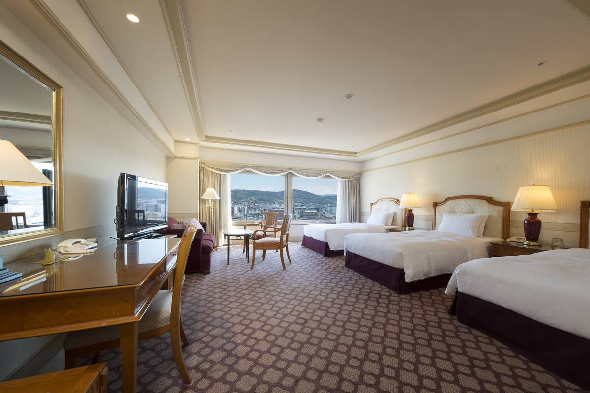 A hotel room with two beds, a desk with a TV, a chair, and a view of the city.