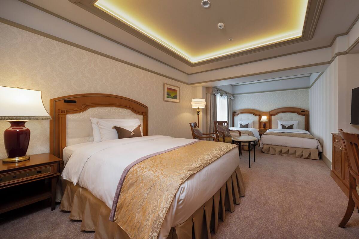 A hotel room with a large bed, two smaller beds, a desk with a TV, and a chair.