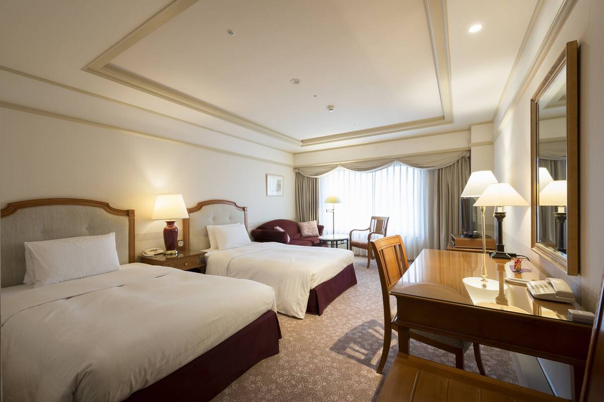 A hotel room with two beds, a desk with a chair, a mirror, and a small table with a lamp.