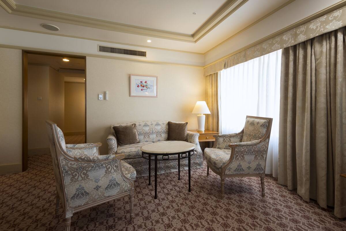 A hotel room with a patterned carpet, a sofa, two armchairs, a small round table, a lamp, and a window with curtains.