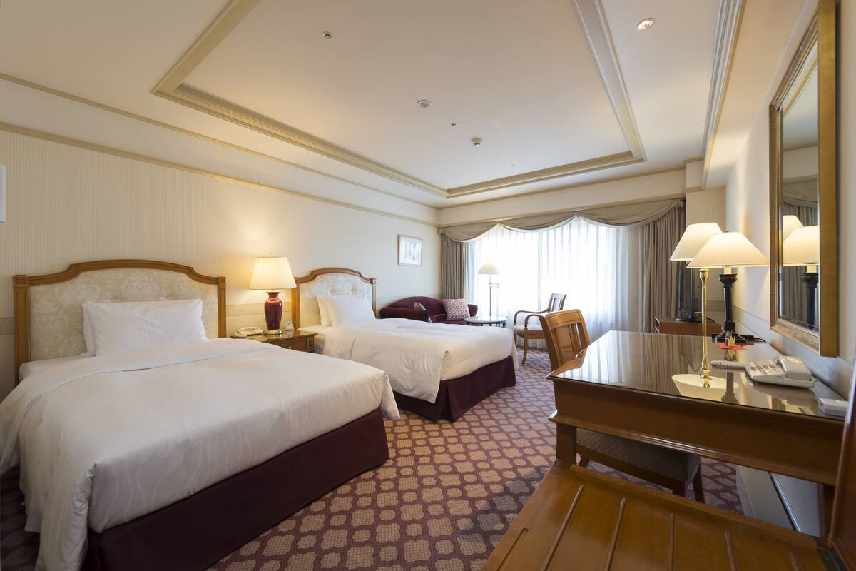 A hotel room with two beds, a desk, a chair, a mirror, and a window with curtains.