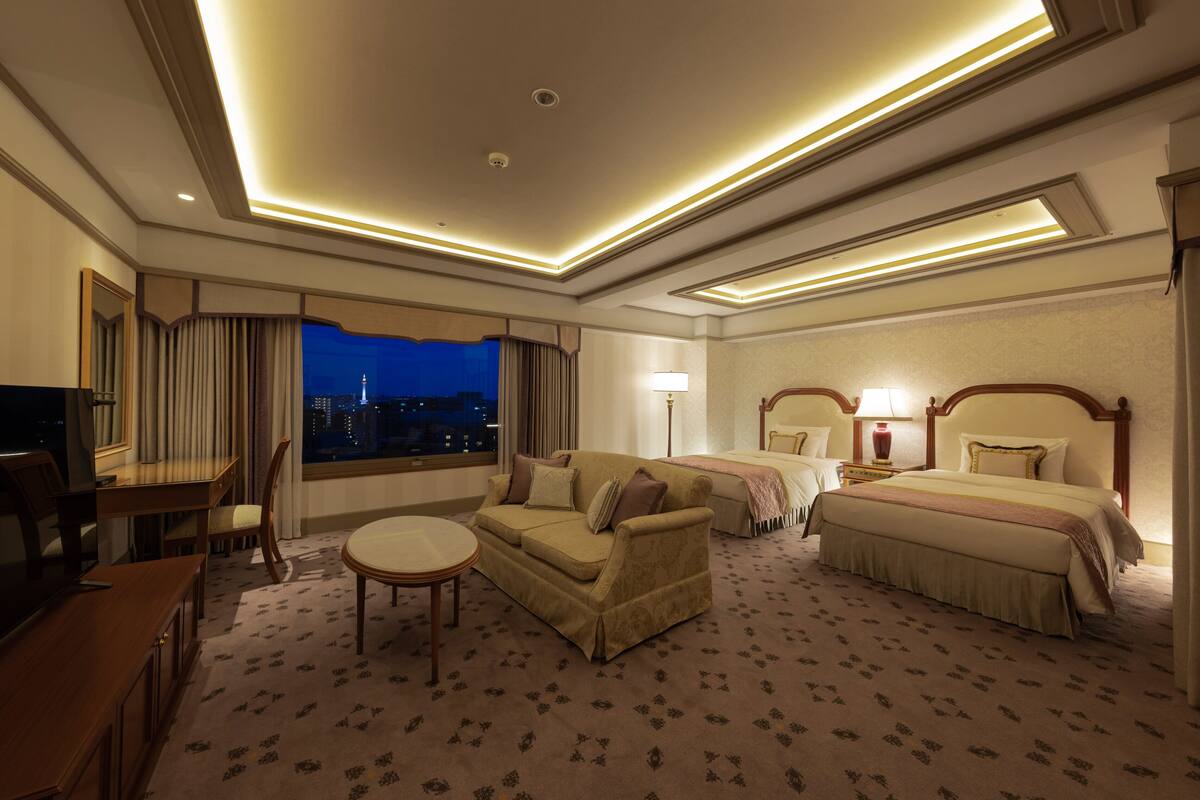 A hotel room with a sofa, two beds, a desk, and a view of the city at night.