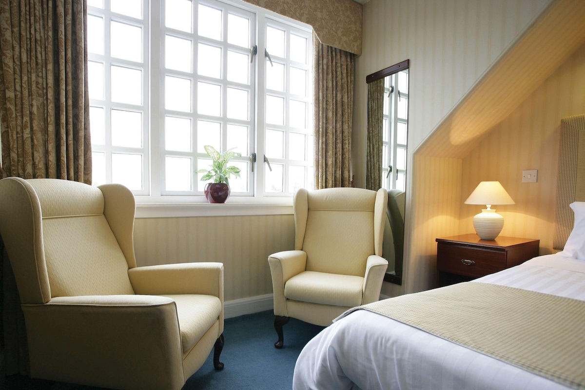 A hotel room with two armchairs, a bed, a nightstand with a lamp, and a window with curtains.