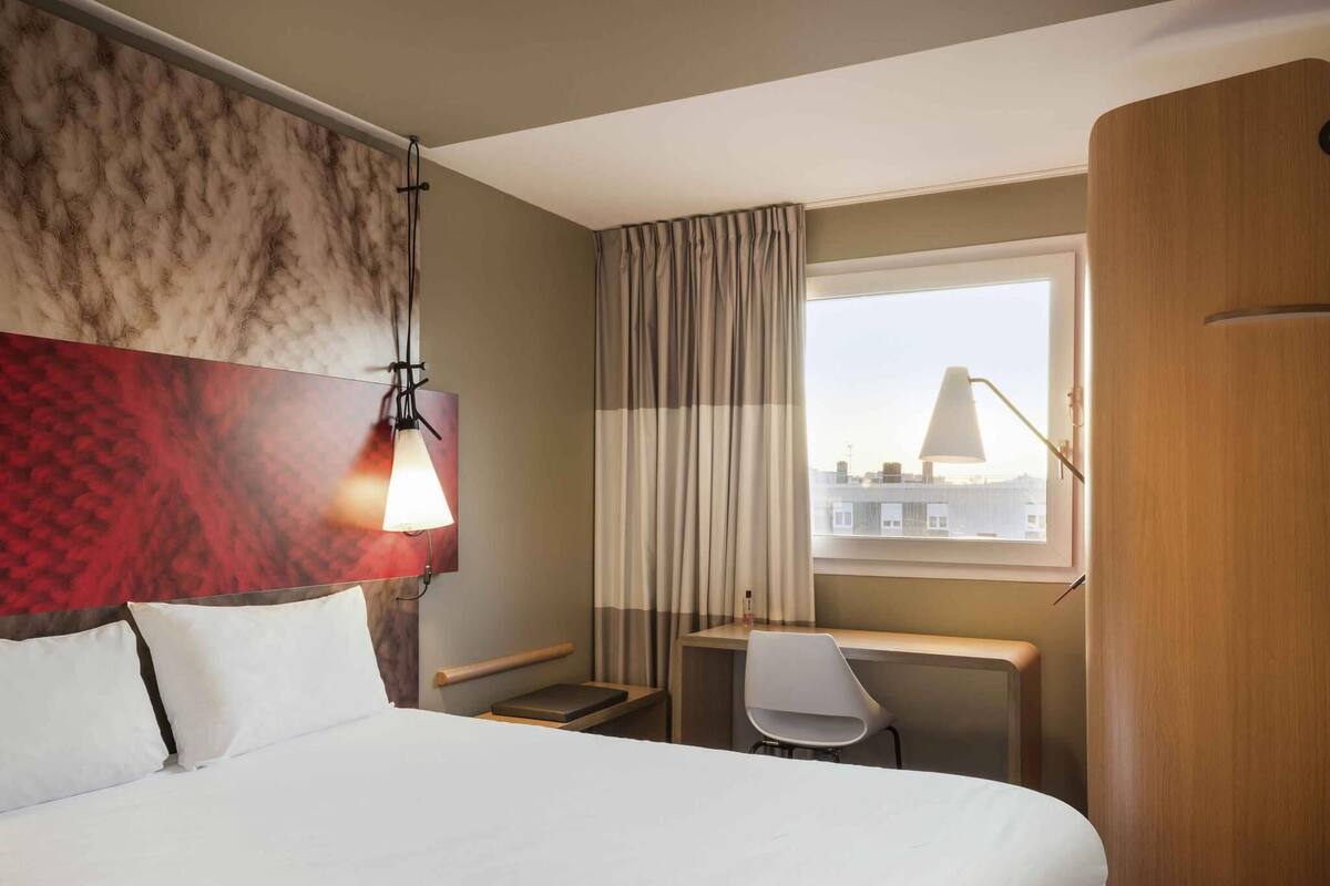 A hotel room with a bed, a desk, a chair, a window with curtains, and a lamp.