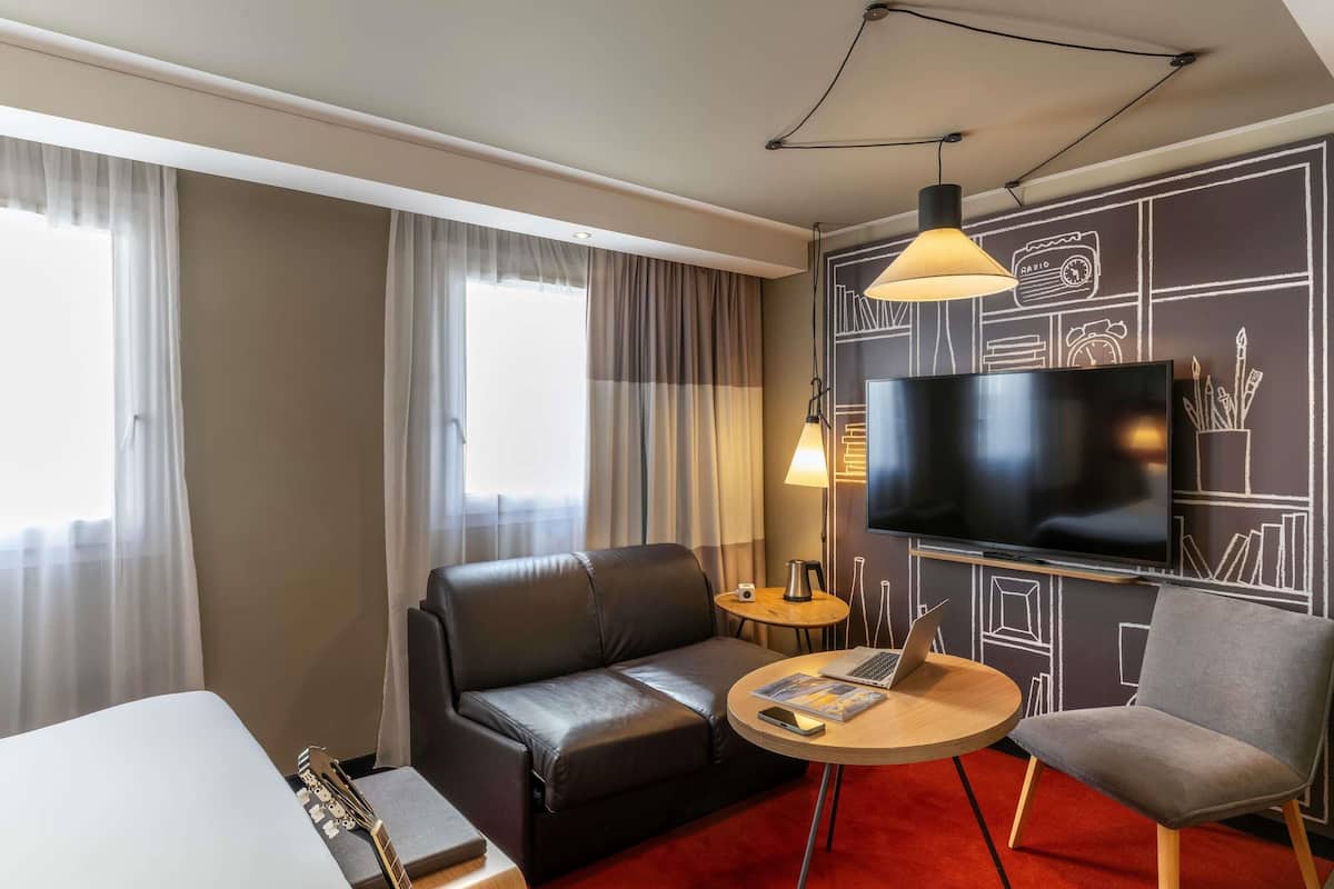 A hotel room with a sofa, a chair, a small table with a laptop, a TV, and a guitar.