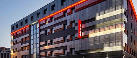 Un moderno edificio hotelero con varios pisos, grandes ventanales y un llamativo letrero rojo que indica que es un hotel ibis.