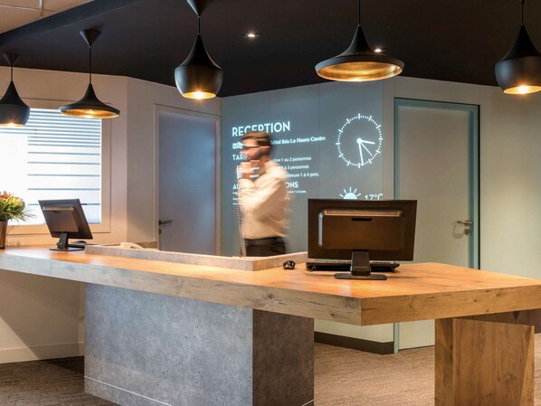 Un mostrador de recepción con una pantalla digital que muestra tarifas de habitaciones y un reloj.