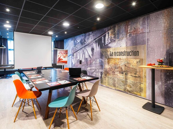 A conference room with a glass table, colorful chairs, a projector screen, and a large mural with the text 'La reconstruction'.