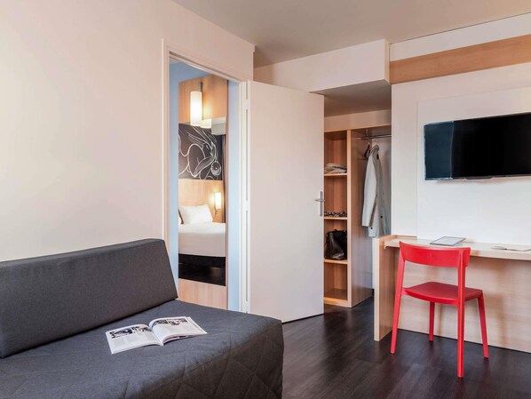 A modern hotel room with a grey sofa, a red chair, a desk, a TV, and a walk-in closet.