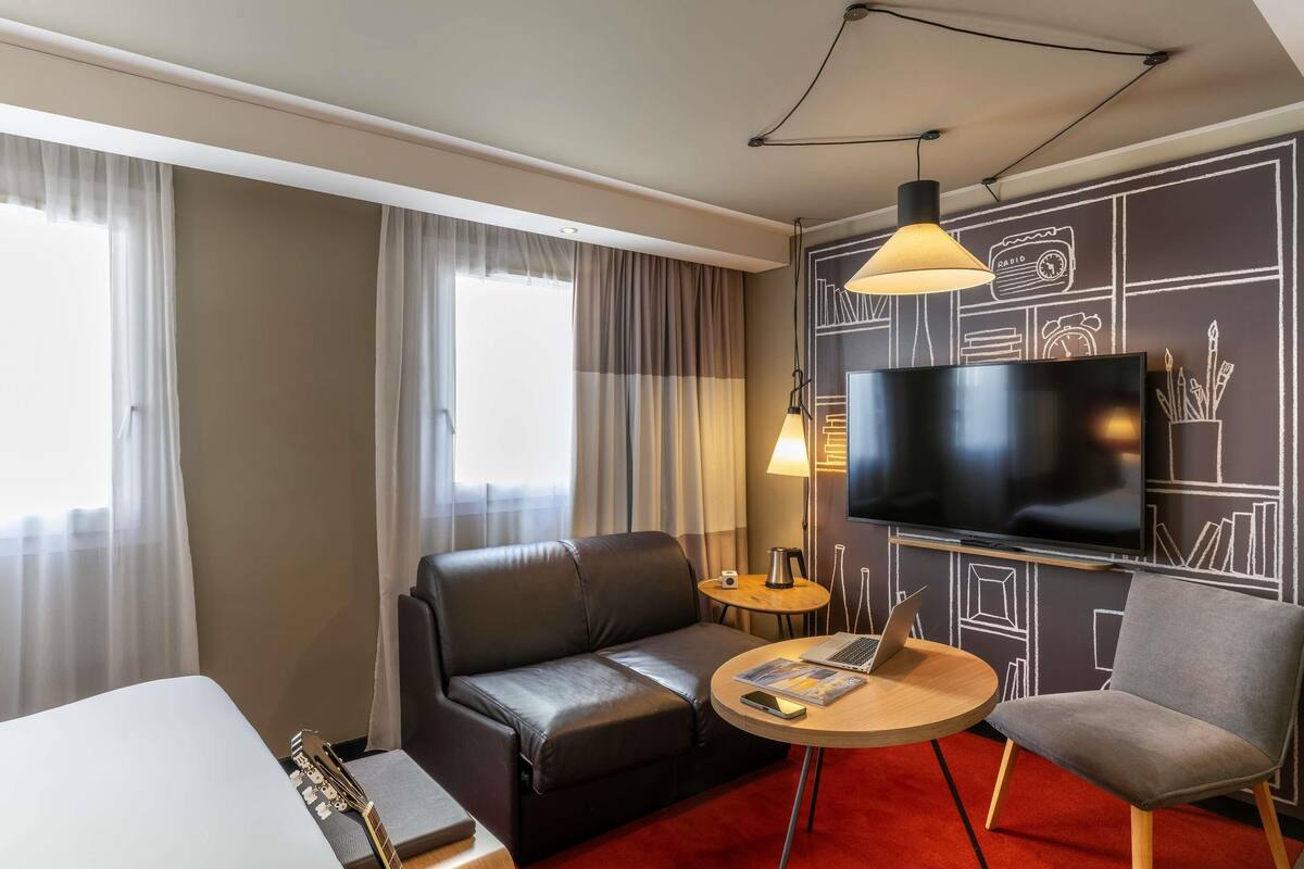 A hotel room with a sofa, a chair, a small table, a TV, and a guitar.