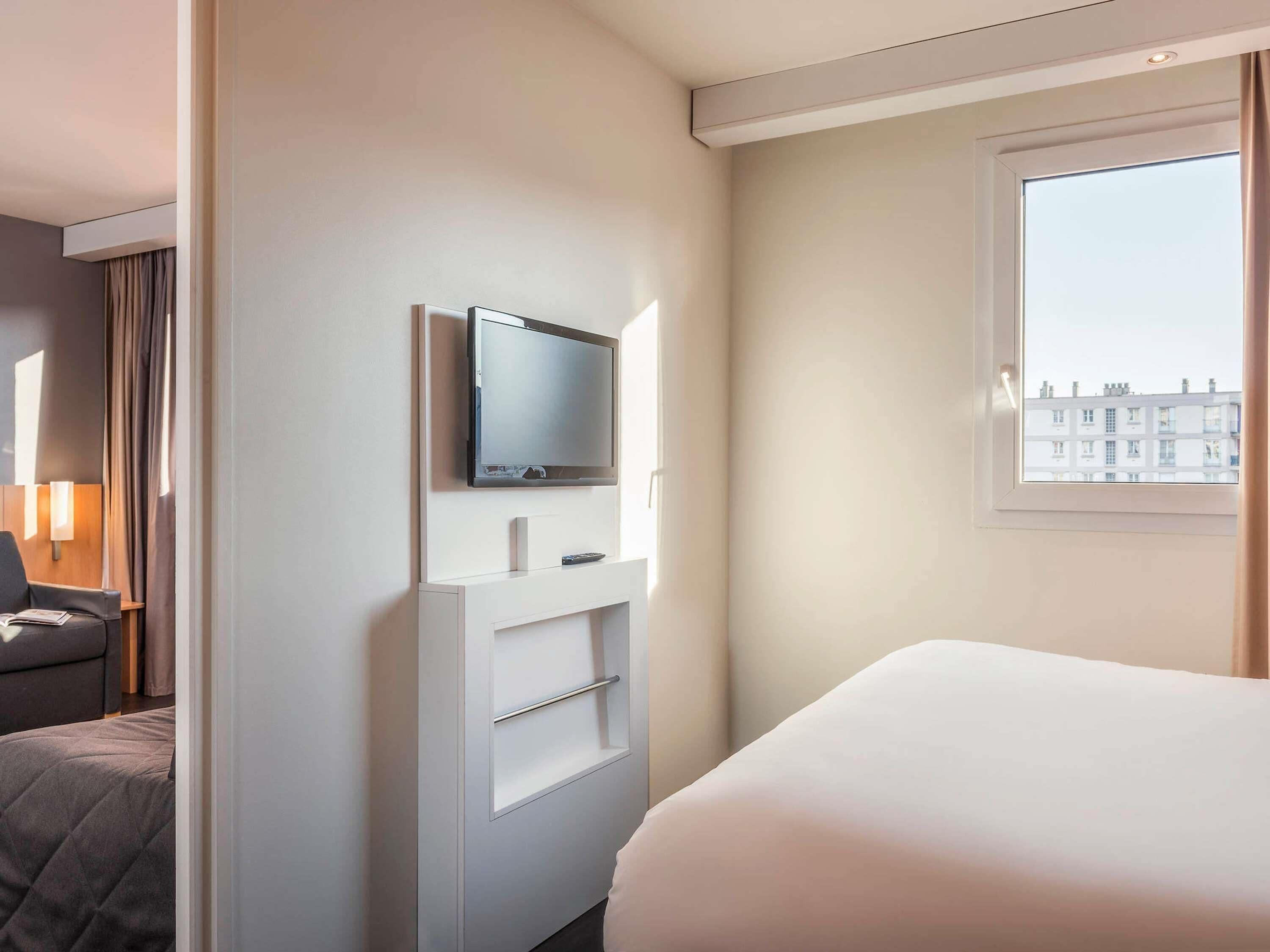 A hotel room with a bed, a TV mounted on a white wall, a window with a city view, and a lamp on a side table.