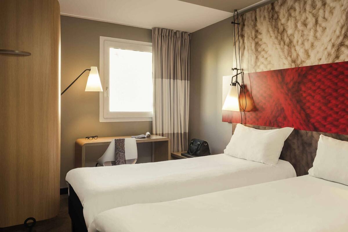 A hotel room with two beds, a desk, a chair, a lamp, a window with curtains, and a headboard with a red pattern.