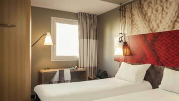 Ein Hotelzimmer mit zwei Betten, einem Schreibtisch, einem Stuhl, einer Lampe, einem Fenster mit Vorhängen und einem roten Muster am Kopfteil.