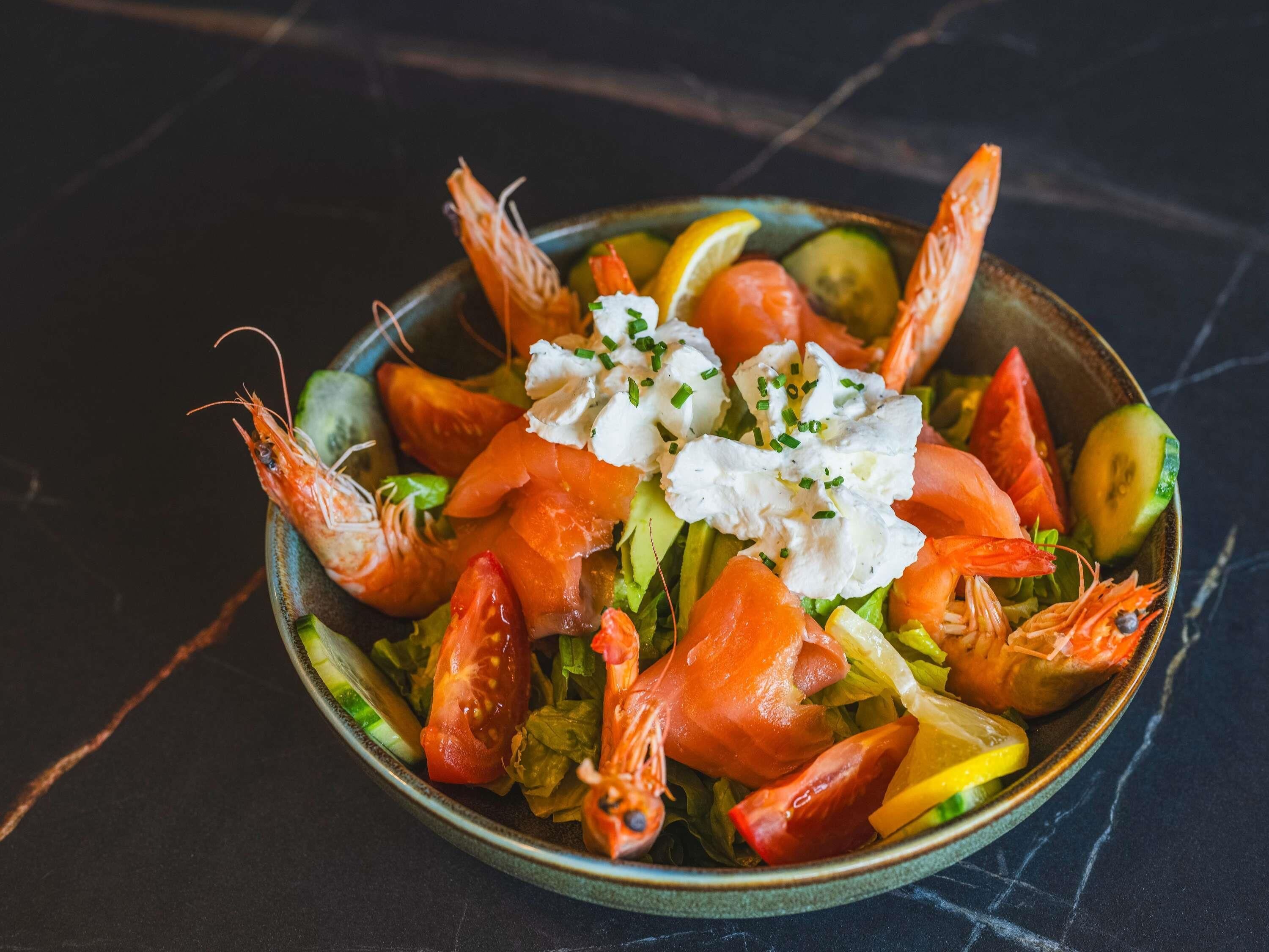 Een schaal vissalade met garnalen, tomaten, komkommers en een lepel room op een donkere ondergrond.