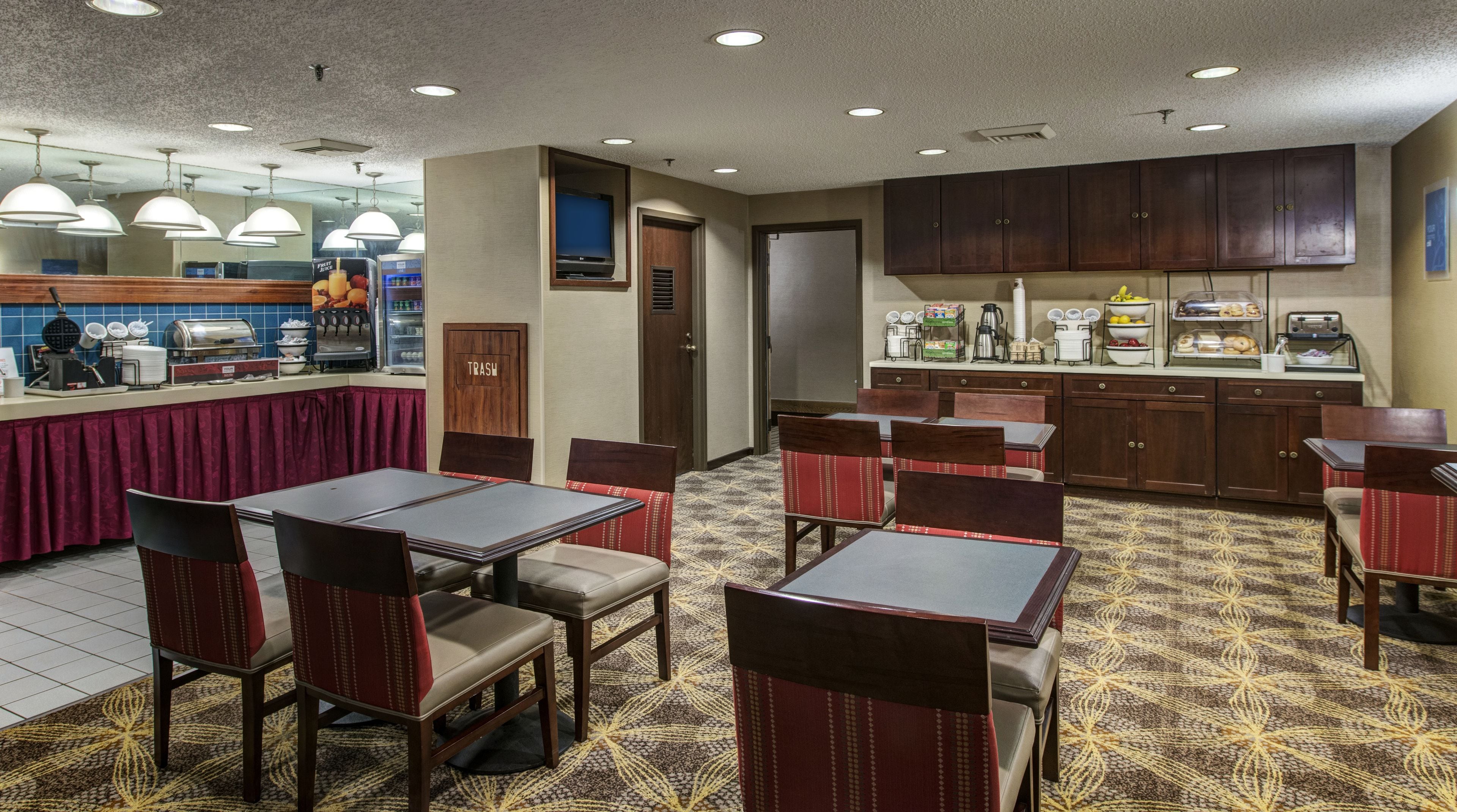 A hotel dining area with tables and chairs, a buffet counter, and a kitchen area.
