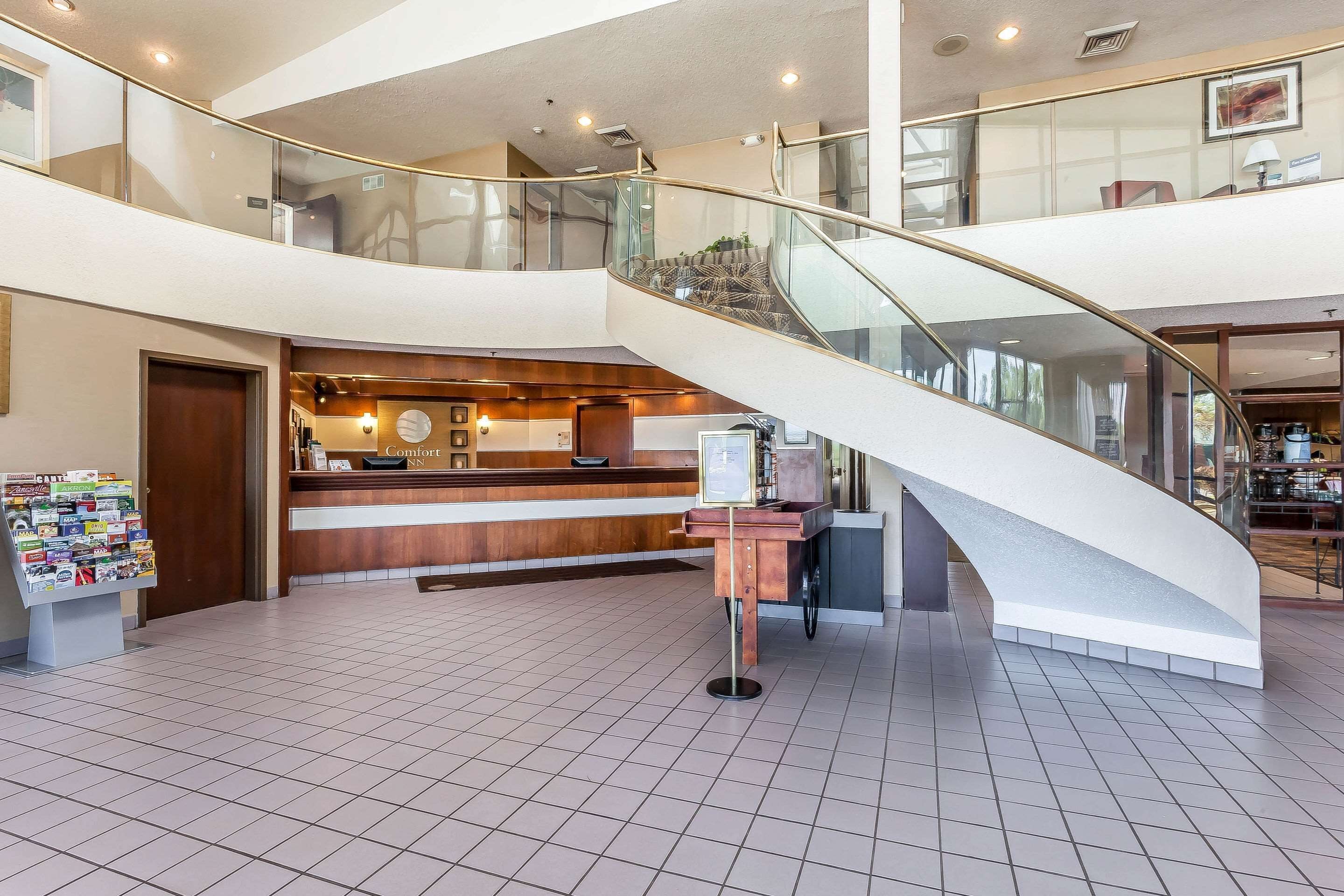 Un vestíbulo de hotel con una escalera de cristal curva, un mostrador de recepción y un tablón de anuncios con varios avisos.
