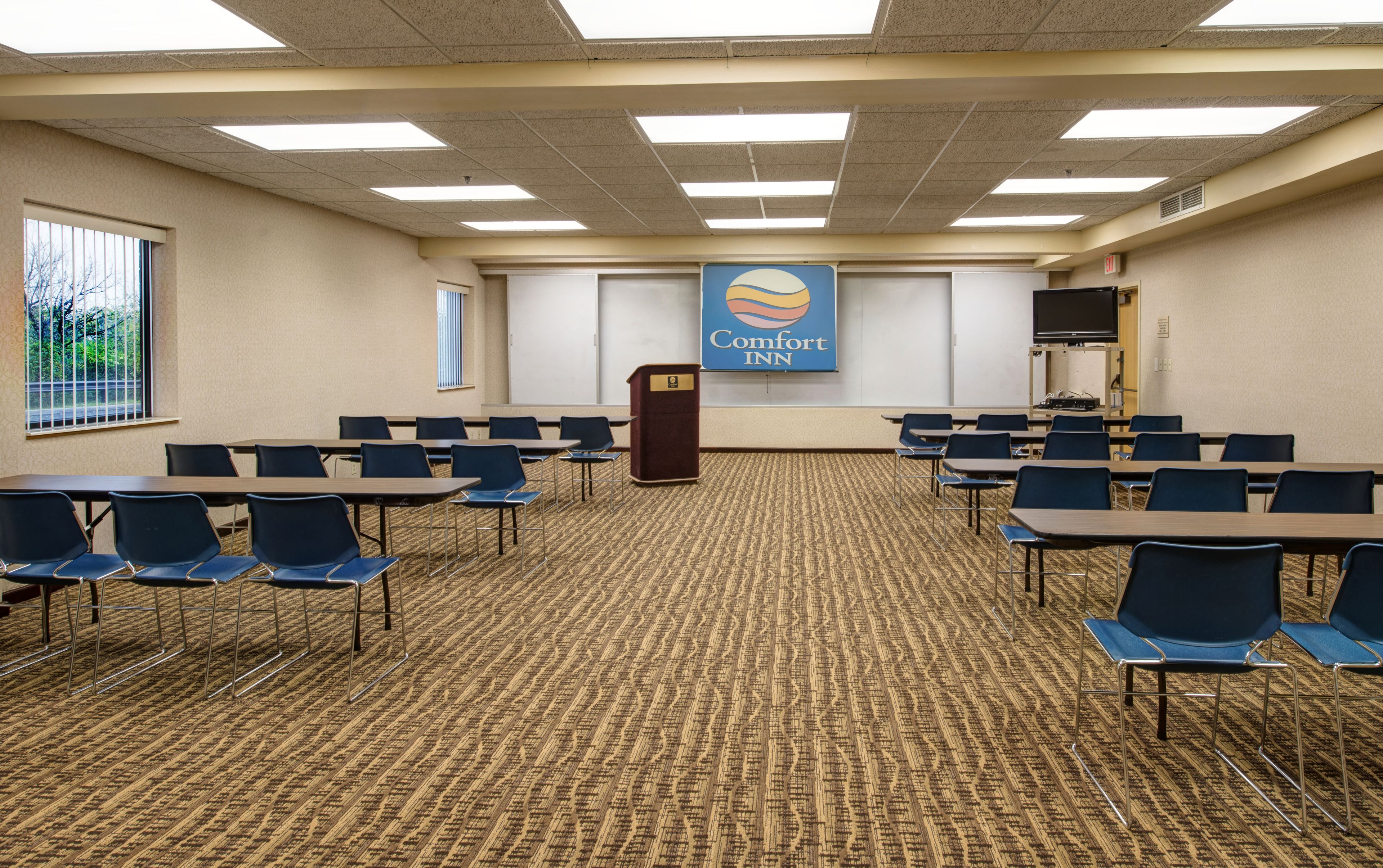 A conference room with a carpeted floor, multiple tables and chairs, a podium, and a Comfort Inn sign on the wall.