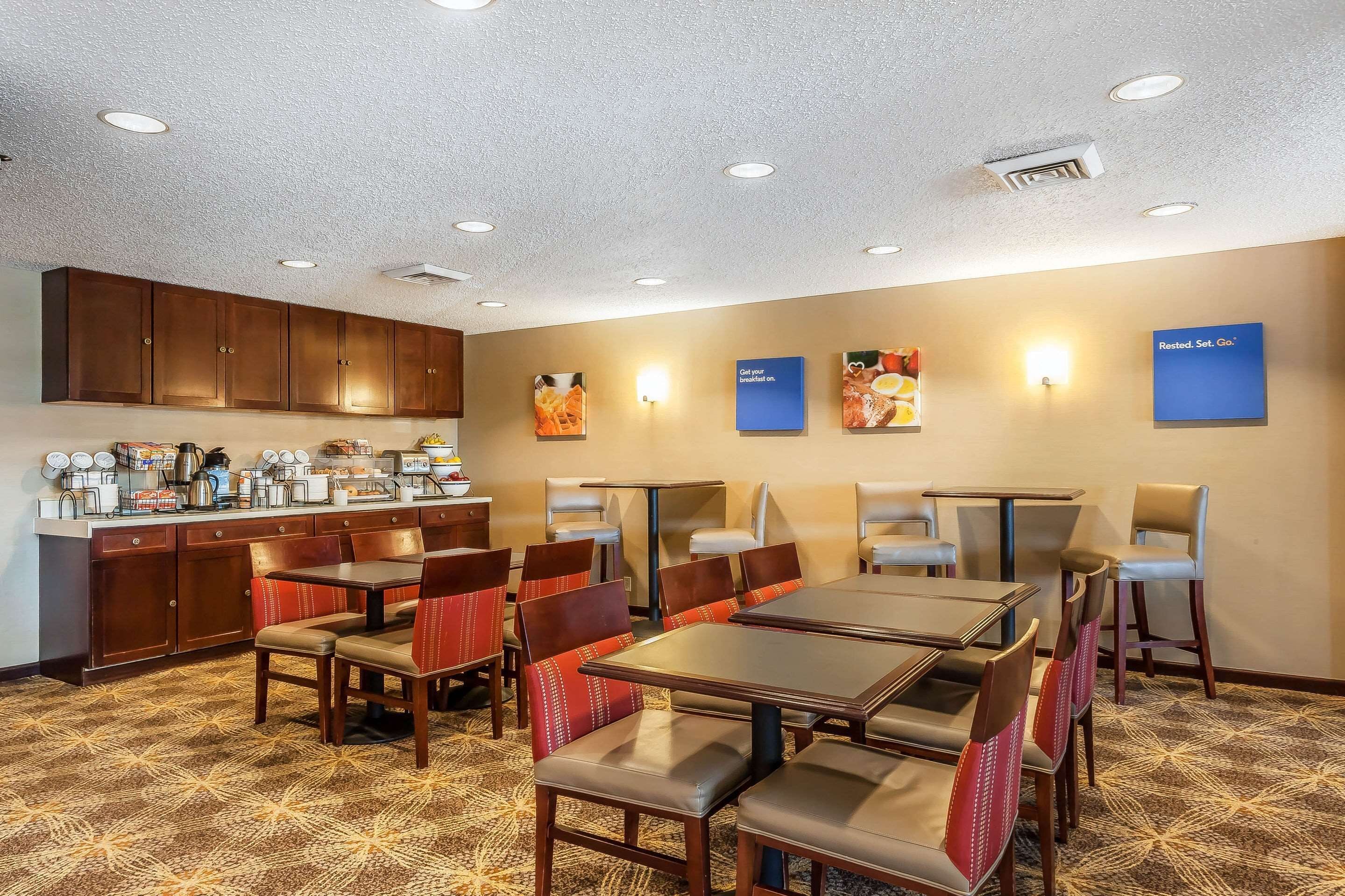 A hotel dining area with multiple tables and chairs, a kitchen counter with coffee machines, and wall-mounted signs.
