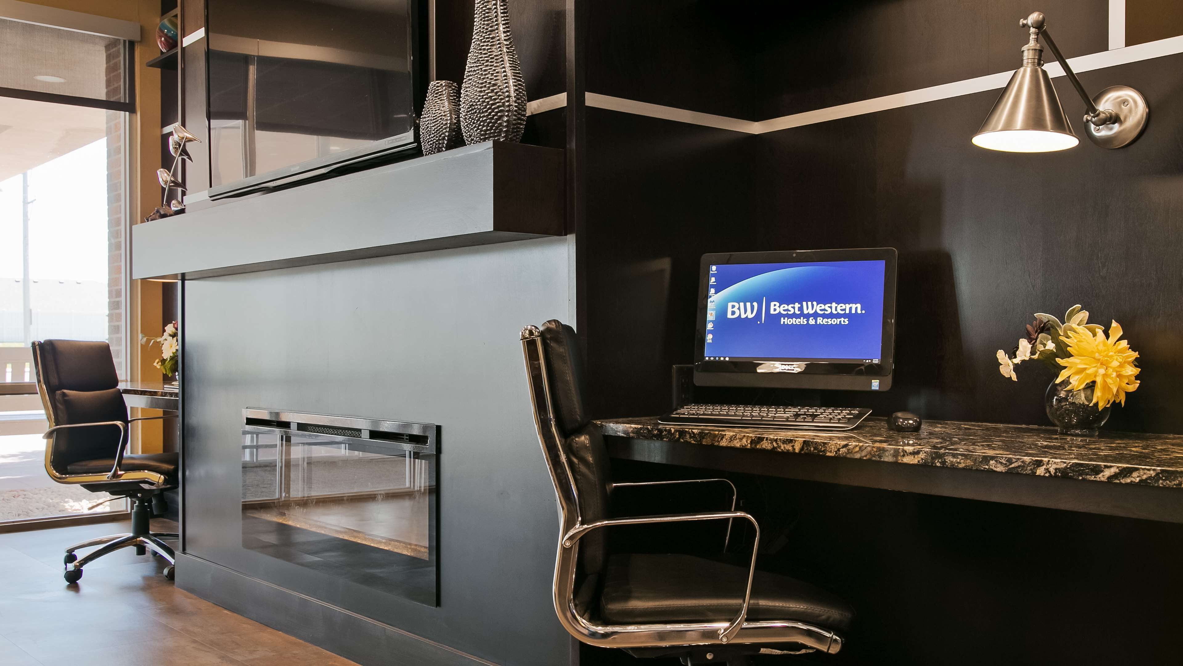 Un moderno mostrador de recepción de hotel con un monitor de computadora mostrando 'BW Best Western. Hotels & Resorts', una silla de cuero negro, un pequeño jarrón con flores amarillas y una lámpara colgante.