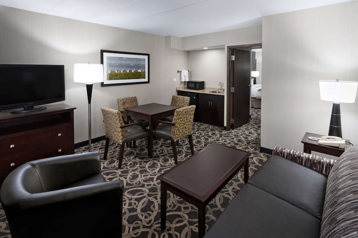 A hotel room with a TV, a dining table with chairs, a sofa, a small table, and a kitchen area with a microwave.