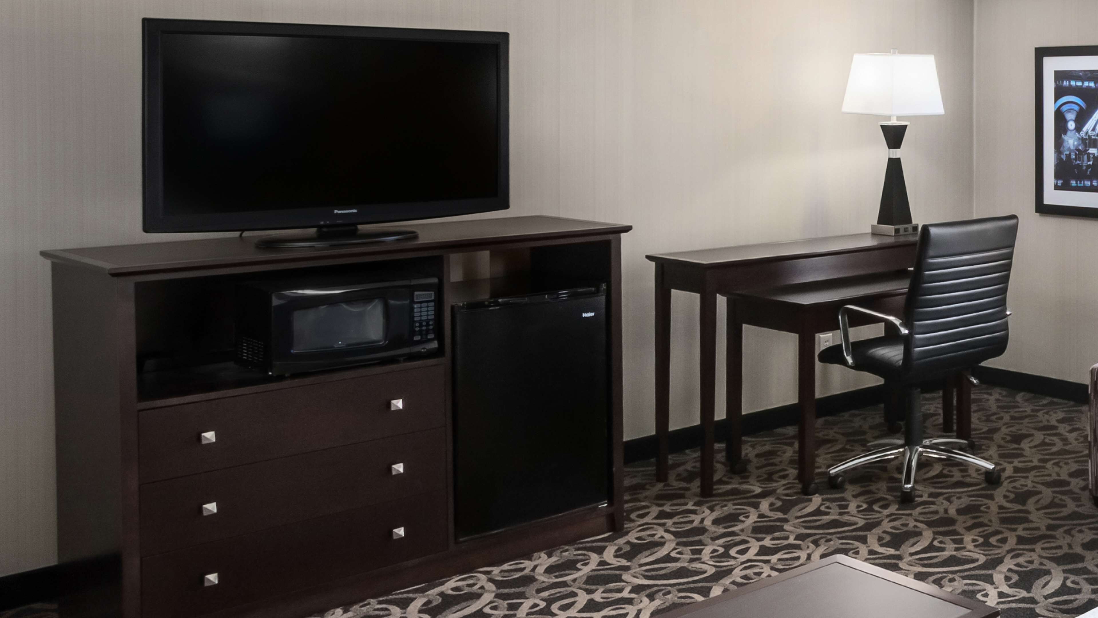 A hotel room with a flat-screen TV, a microwave, a mini-fridge, a desk, a chair, a lamp, and a patterned carpet.