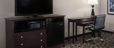 A hotel room with a flat-screen TV, a microwave, a mini-fridge, a desk, a chair, a lamp, and a patterned carpet.