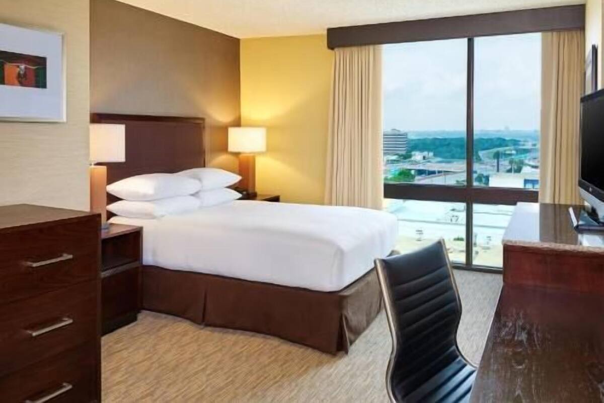 Une chambre d’hôtel équipée d’un lit, de tables de chevet, d’un bureau, d’une chaise et d’une grande fenêtre donnant sur un paysage agréable.