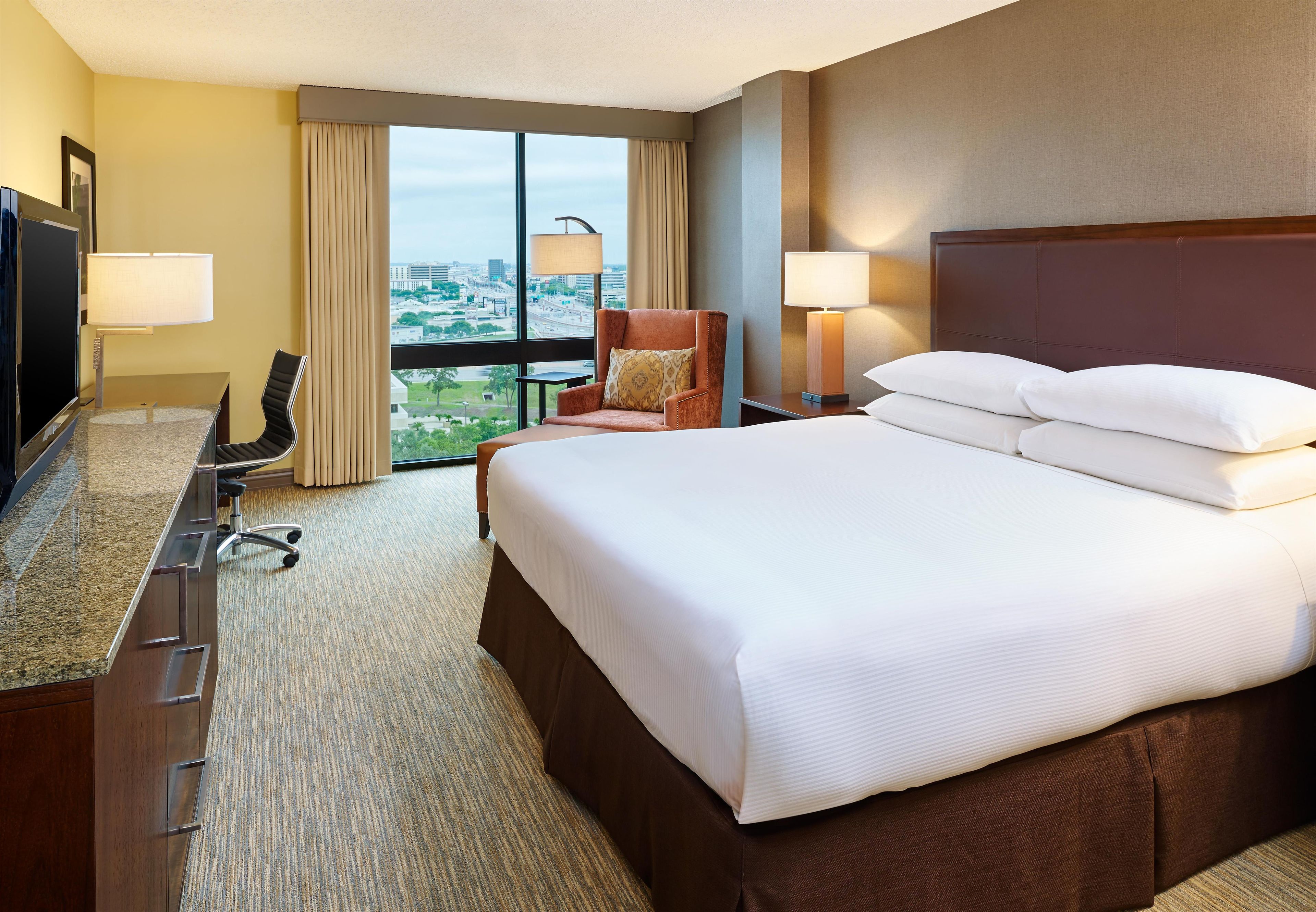 Habitación de hotel con una cama grande, un escritorio con televisor, una silla, dos lámparas y vistas a la ciudad a través de amplios ventanales.