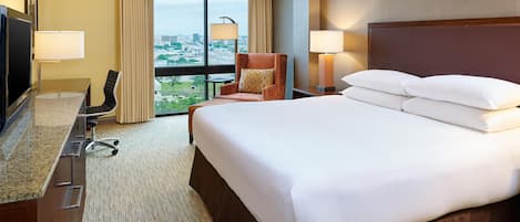 Habitación de hotel con una cama grande, un escritorio con televisor, una silla, dos lámparas y vistas a la ciudad a través de amplios ventanales.