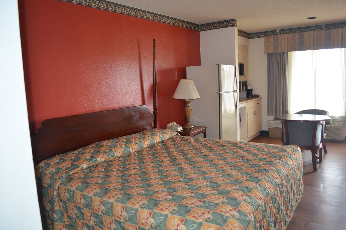 A hotel room with a large bed, a nightstand with a lamp, a small table, and a kitchenette area.