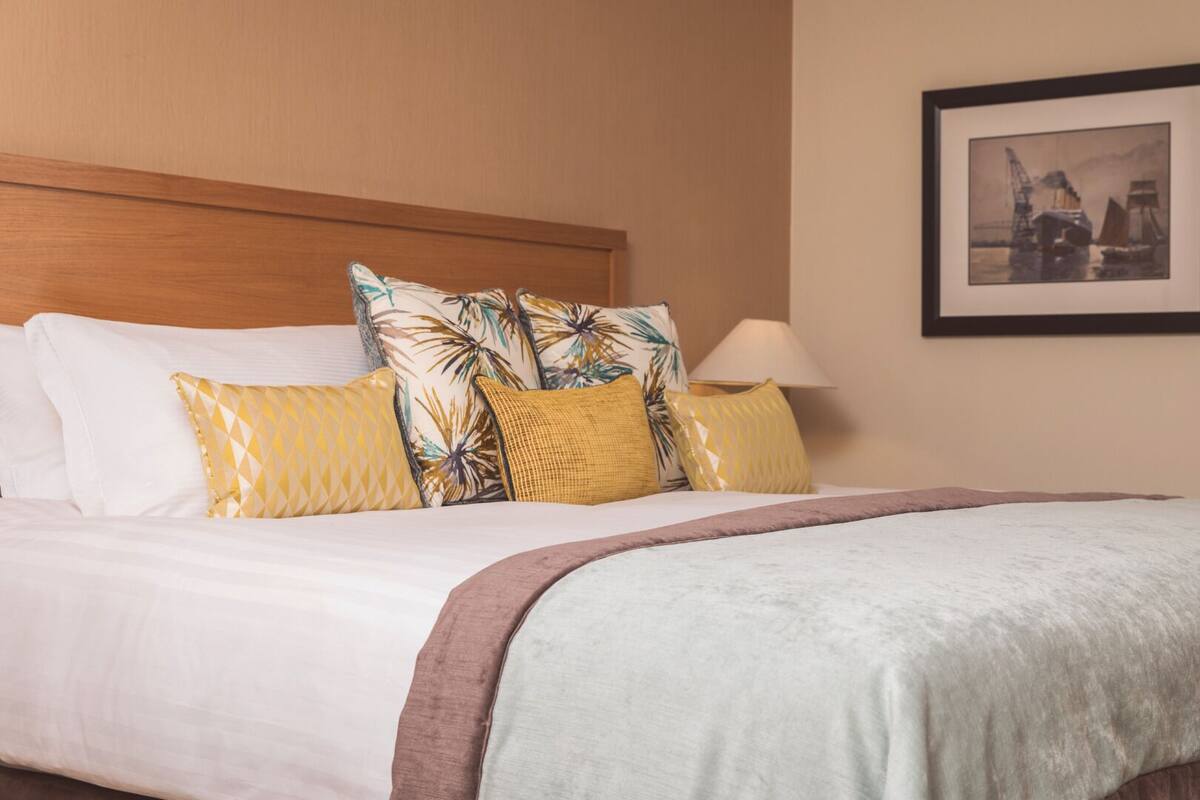 Una cama bien hecha con sábanas blancas, un cabecero marrón y almohadones decorativos en amarillo y blanco.