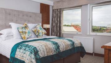 A hotel room with a bed, a window with a view of buildings, a radiator, and a wooden nightstand.