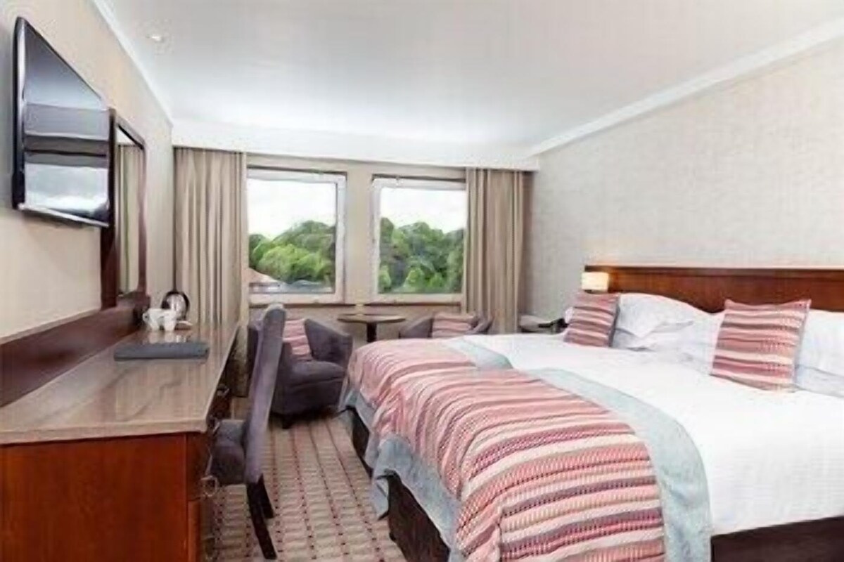 A hotel room with a bed, a desk, a chair, and a view of greenery through the windows.