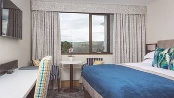Quarto de hotel com cama, escrivaninha, cadeira, mesinha e janela com cortinas.