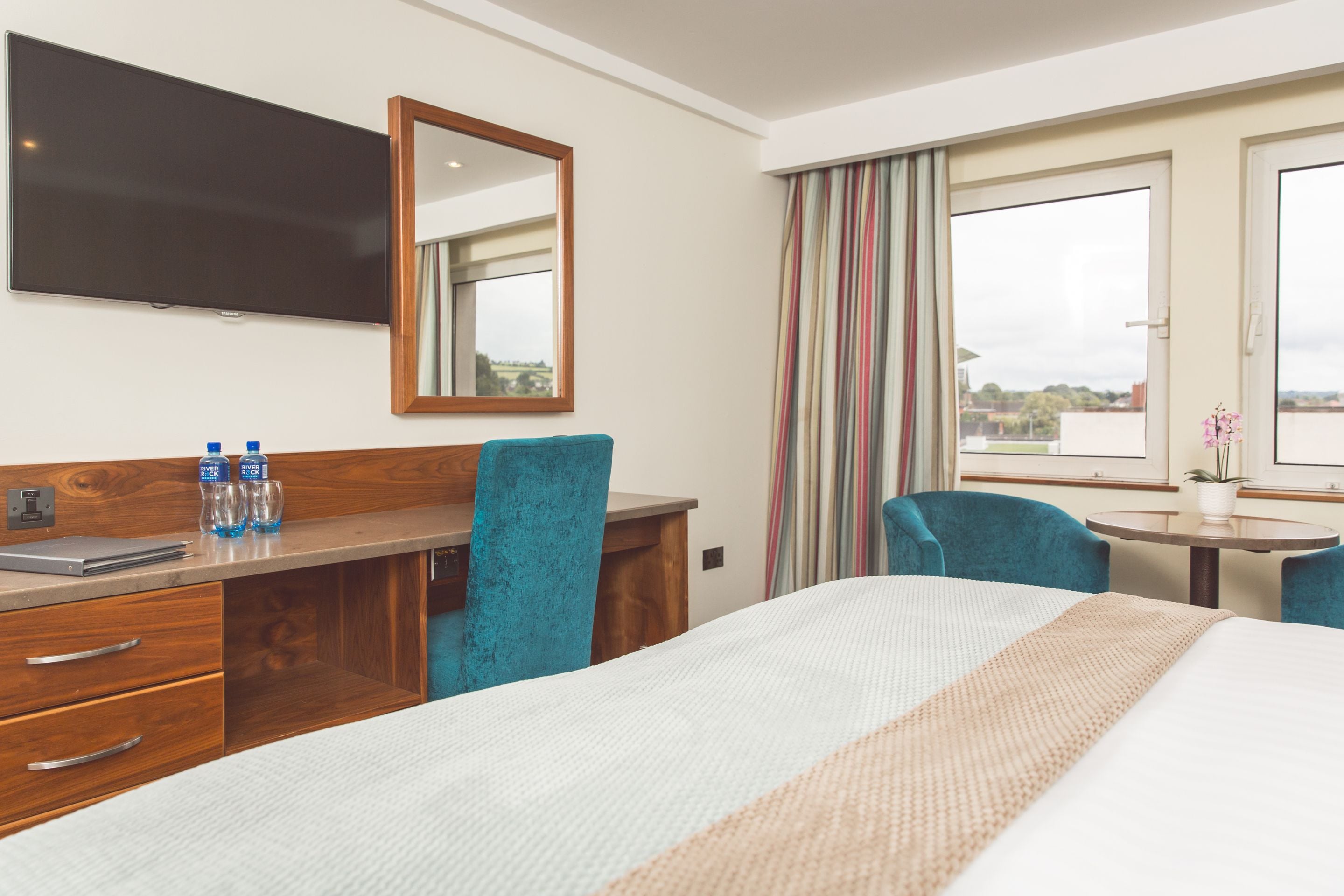 A hotel room with a bed, a desk with a laptop, a mirror, a TV, a window with curtains, and a small table with a plant.