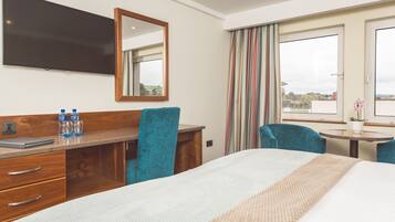 A hotel room with a bed, a desk with a laptop, a mirror, a TV, a window with curtains, and a small table with a plant.