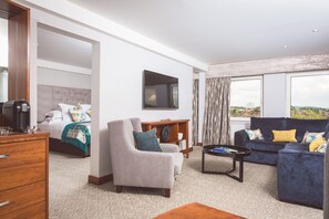 Habitación de hotel con cama, zona de estar con sofá y sillón, una mesa de centro y un amplio ventanal con vistas a la ciudad.