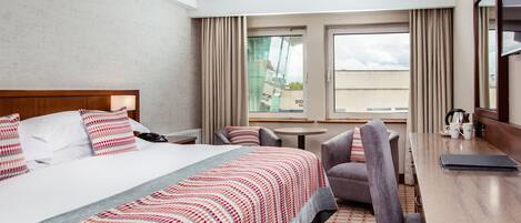 A hotel room with a large bed, a desk, a chair, a small table, and a window with curtains.