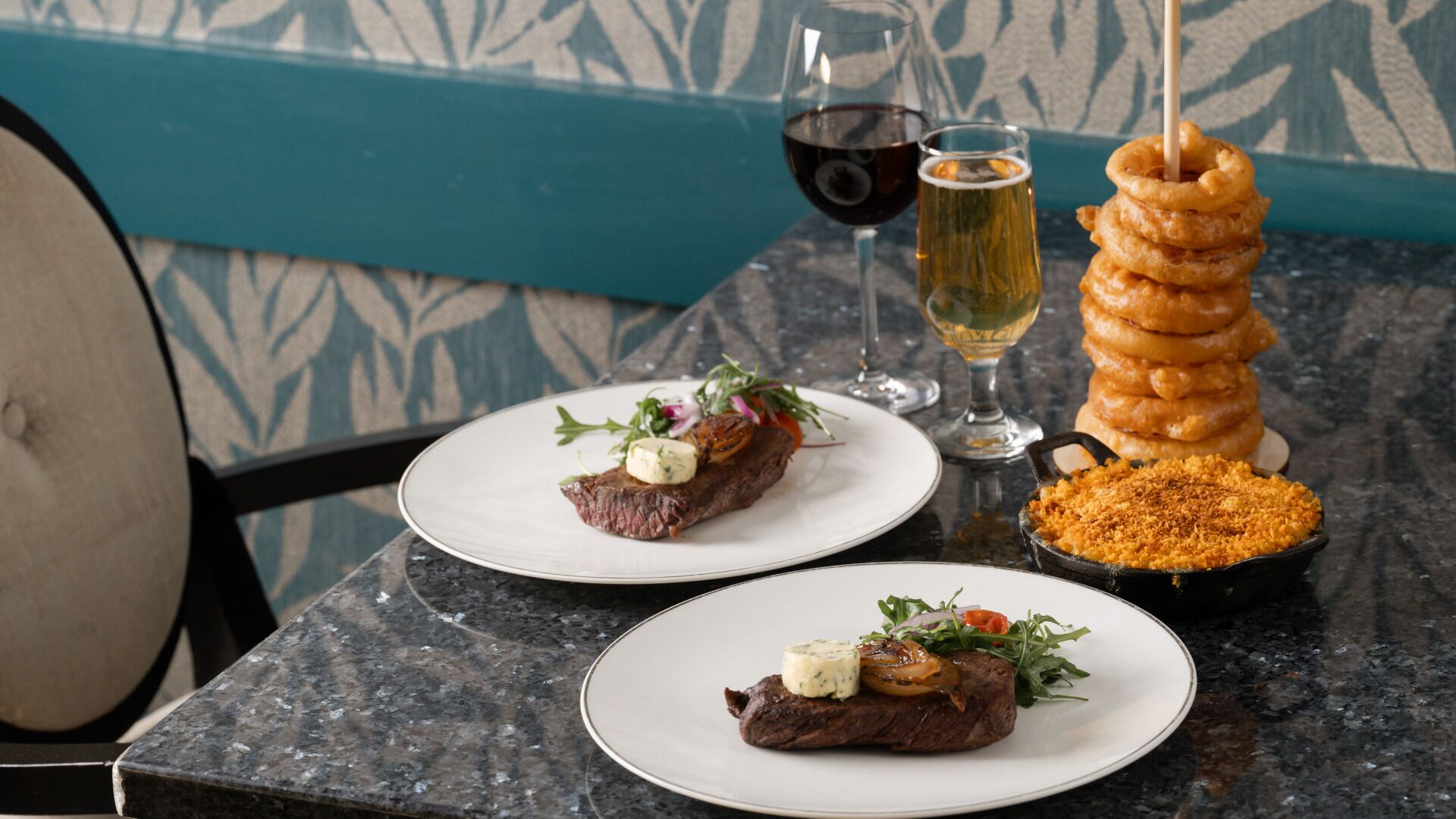 Two plates of steak with vegetables and a side of breaded dish, a glass of red wine, and a glass of white wine on a table.