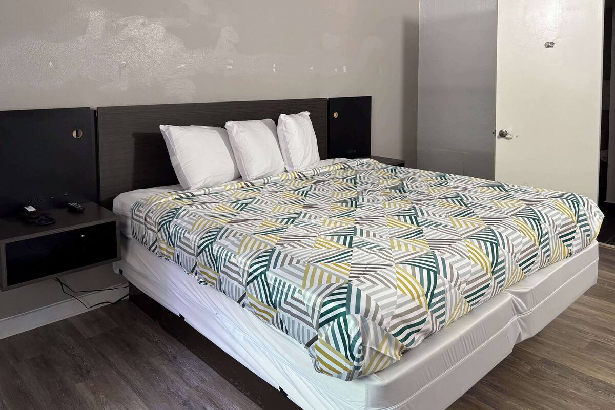 A neatly made bed with a patterned bedspread, a dark headboard, and a bedside table with a phone and charger.