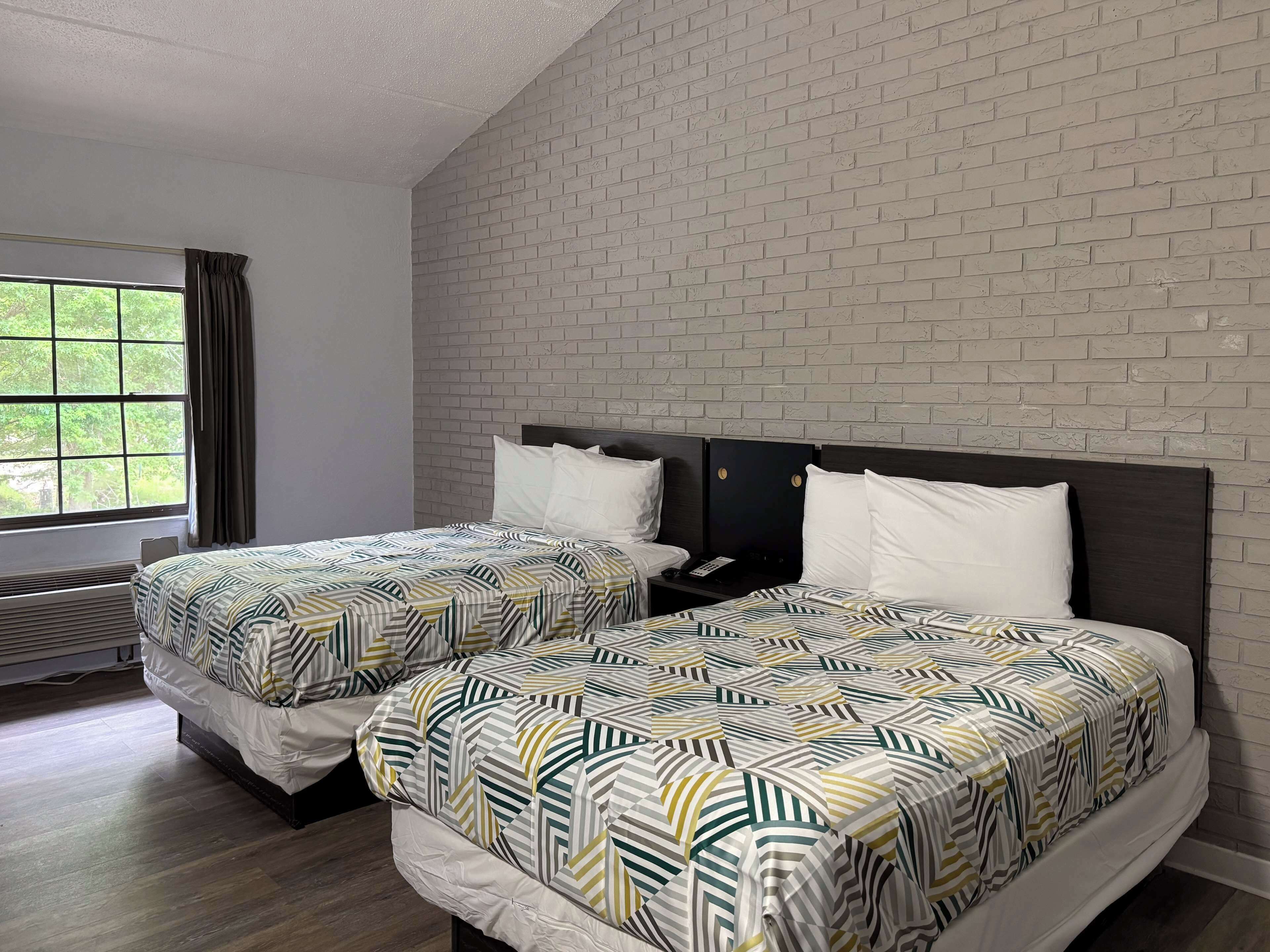 A hotel room with two beds, a brick-patterned wall, a window with curtains, and a wooden floor.