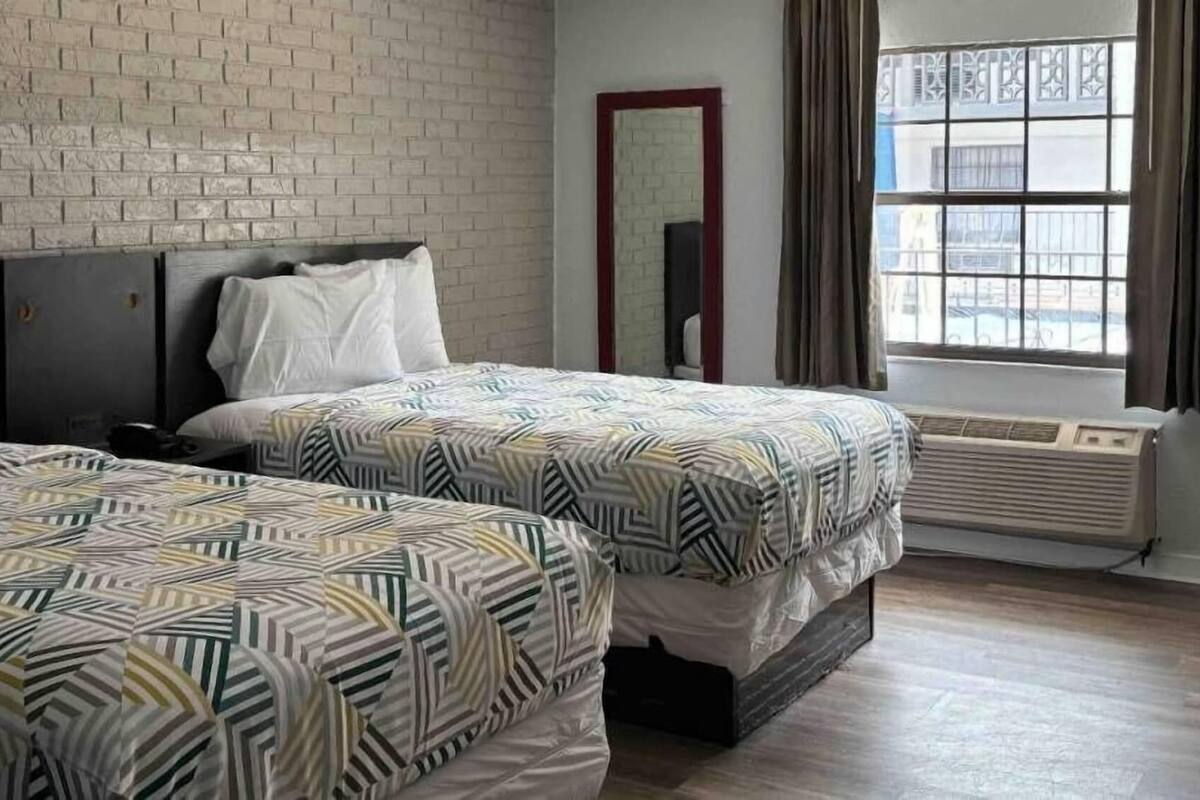 A hotel room with two beds, a brick wall, a large window with curtains, and a mirror.