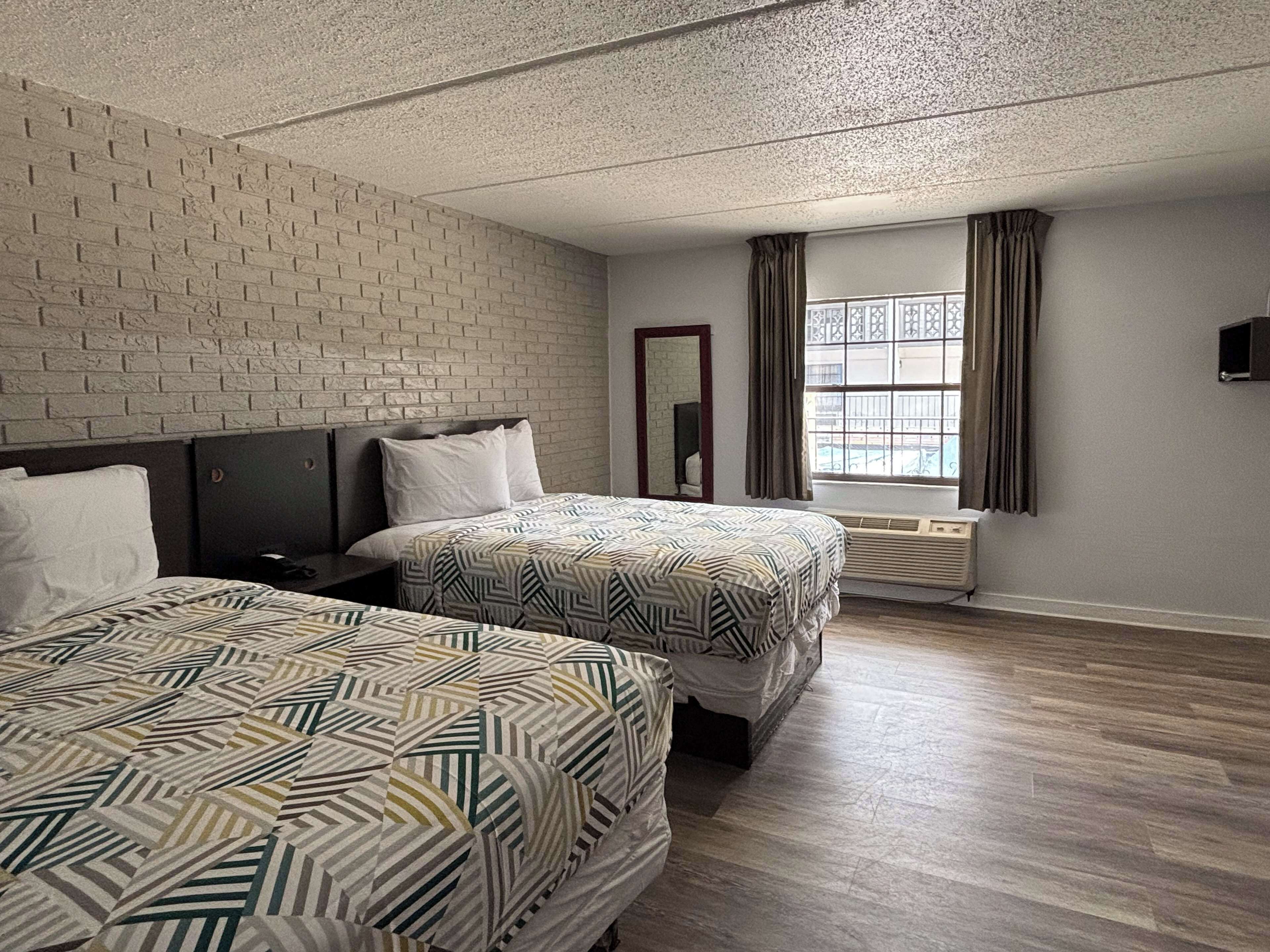 A hotel room with two beds, a brick accent wall, a large window with curtains, and a flat-screen TV mounted on the wall.