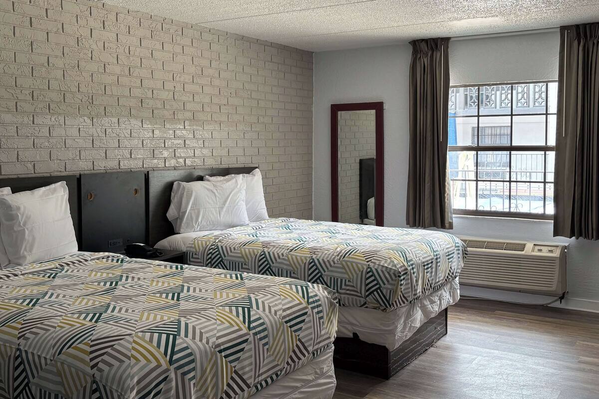 A hotel room with two beds, a brick wall, a window with curtains, and a mirror.