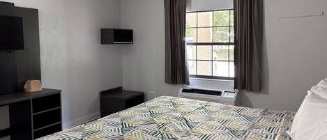 A hotel room with a bed, a TV mounted on the wall, a window with curtains, and a small table with a telephone.